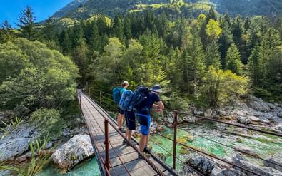 Drie wandelaars met rugzakken steken een houten voetbrug over een turquoise rivier in Lepena, Julische Alpen. Dicht bos en bergen op de achtergrond.