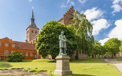 Bronzen standbeeld van Hans Christian Andersen op wit voetstuk in park met kerktoren en historische bakstenen gebouwen in Odense, Denemarken.