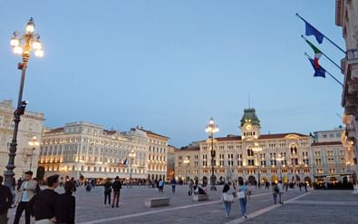 Piazza Unità d'Italia in Triëst bij schemering met verlichte historische gebouwen, sierlijke straatlantaarns en mensen die over het plein lopen.