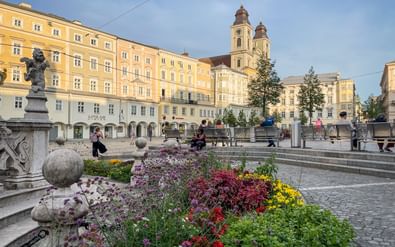 Linz Hauptplatz met kleurrijke bloembedden op de voorgrond, gele historische gebouwen en kerk met twee torens onder blauwe lucht.