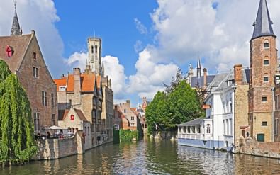 Gracht in Brugge met middeleeuwse bakstenen gebouwen, kerktorens en het beroemde Belfort zichtbaar op de achtergrond onder blauwe lucht.
