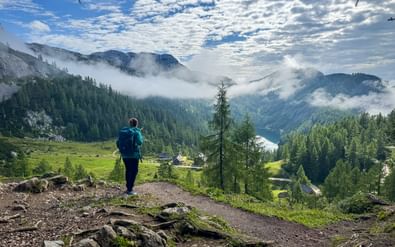Wandelaar met blauwe rugzak op bergpad bij Tauplitzalm, uitkijkend over het Steirersee-meer omringd door beboste bergen en ochtendmist.