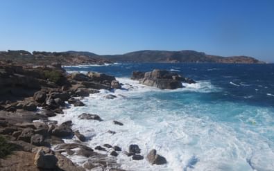 Rotsachtige kustlijn bij Revellata, Corsica met turquoise golven die tegen donkere rotsen slaan. Heuvels en landtongen zichtbaar in de verte onder heldere lucht.