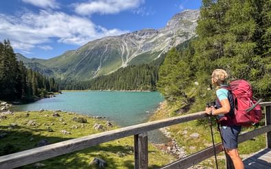 Wandelaar met rode rugzak en wandelstokken bij houten hek met uitzicht op turquoise bergmeer omringd door beboste bergen.