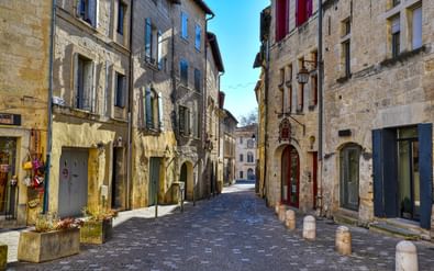 Smalle geplaveide straat in Uzès, Provence, omzoomd met oude stenen gebouwen met blauwe en rode luiken onder een heldere blauwe lucht.