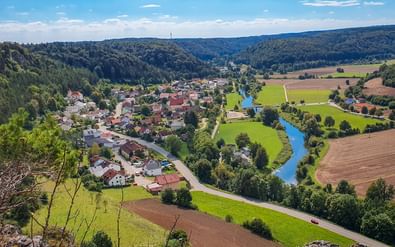 Luchtfoto van het dorp Arnsberg in het Altmühltal met blauwe rivier die door groene weiden en velden kronkelt, omringd door beboste heuvels.