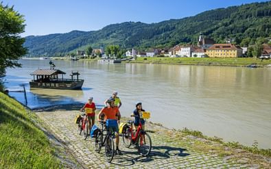 Groep fietsers met fietsen op geplaveide rivieroever kijkt naar veerboot op Donau. Oostenrijks dorp Engelhartszell met heuvels op achtergrond.