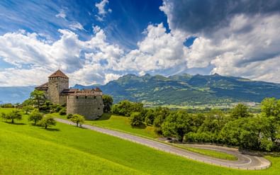 Kasteel Vaduz met Alpenlandschap Kasteel Vaduz op een groene heuvel in Liechtenstein met kronkelende weg, omgeven door weelderige weiden en Alpengebergte.