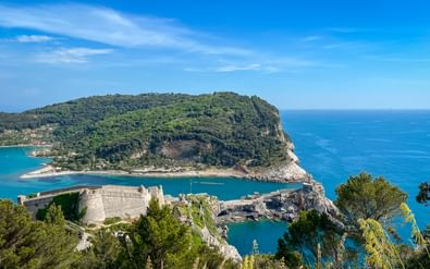 Luchtfoto van Porto Venere fort op rotsachtig schiereiland met turquoise water. Groen bebost eiland verbonden door dam onder blauwe lucht.