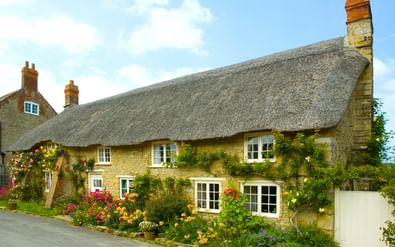 Traditioneel rieten cottage met honingkleurige stenen muren in Abbotsbury, Dorset. Kleurrijke bloemen in de voortuin onder een blauwe lucht.