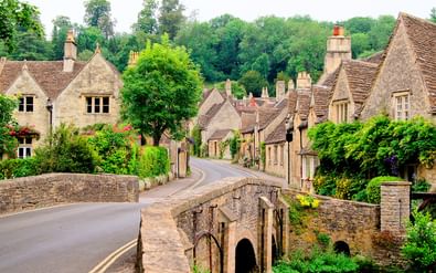Cotswold Way dorp met stenen brug Historisch Cotswold dorp met honingkleurige stenen huizen en middeleeuwse brug. Weg kronkelt door dorp met groene bomen en tuinen.