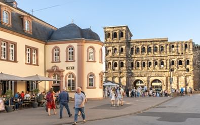 Oude Romeinse Porta Nigra poort in Trier met toeristen op het plein. Historische barokgebouwen met café-terras links zichtbaar.