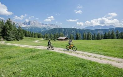 Twee fietsers op een grindpad door een groene alpenweide in de Dolomieten, met bossen, een houten hut en bergtoppen onder blauwe lucht.