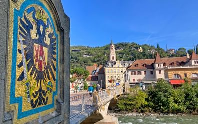 Brug in Merano met mozaïek adelaarsembleem, over groene rivier. Historische stad met kerktoren en gebouwen op heuvel.