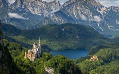 Kasteel Neuschwanstein op een beboste heuvel met dramatische Alpenbergen en een blauw meer op de achtergrond onder bewolkte luchten.