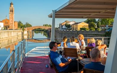 Passagiers ontspannen op het zonnedek van MS Ave Maria in Portegrandi, met kanaal, brug en kerktoren op de achtergrond.