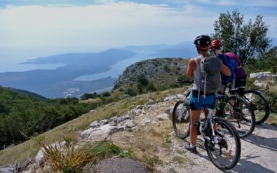 Twee fietsers met rugzakken staan met mountainbikes op een rotsachtig bergpad, uitkijkend over de Baai van Kotor in Montenegro.