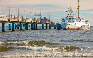 Wit-oranje veerboot aangemeerd aan de pier van Bansin op Usedom. De pier heeft blauwe pilaren en strekt zich uit in de Oostzee met golven op voorgrond.