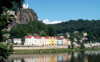 Kleurrijke historische gebouwen langs de rivieroever in Decin, Tsjechië, met wit kasteel op rotsklif erboven en beboste heuvels op achtergrond.