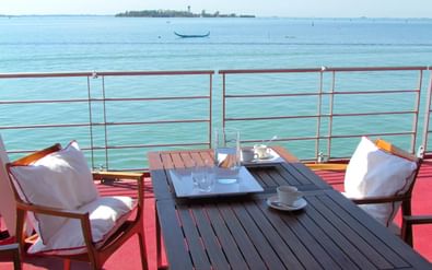 Zonnedek met houten tafel, stoelen met witte kussens en rode vloer met uitzicht op de Venetiaanse lagune met verre eilanden.