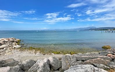 Rotsachtige oever van het Gardameer bij Bardolino met helder turquoise water, stenen golfbreker en bergen in de verte onder blauwe lucht.