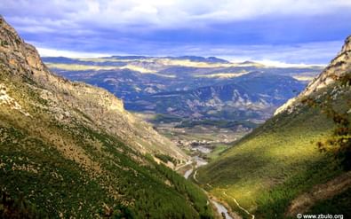Breed uitzicht op Hidden Valley, Albanië, met steile berghellingen, kronkelende rivier en verre bergketens onder bewolkte luchten.