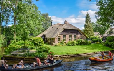 Traditioneel rietgedekt boerderijtje met roze muren aan een gracht in Giethoorn. Toeristen in bootjes varen langs het groene gazon en weelderige bomen.