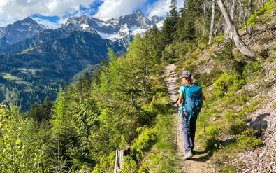 Vrouwelijke wandelaar met rugzak en stokken op bergpad in Hollersbachtal, Hohe Tauern. Besneeuwde toppen en groene bossen zichtbaar op achtergrond.
