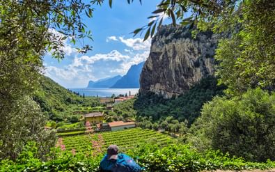 Persoon zittend op stenen muur met uitzicht op wijngaardrijen met dramatische rotswand en Gardameer op de achtergrond.