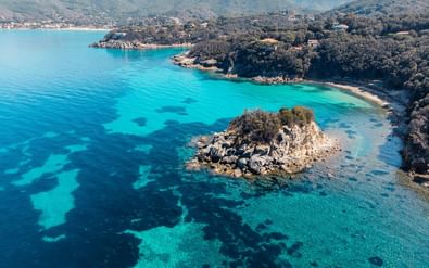 Luchtfoto van Elba's kust met kristalhelder turquoise water, een klein rotseiland, beboste heuvels en verspreide gebouwen langs de baai.
