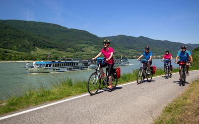 Vier fietsers op Donaufietspad met riviercruiseschip MS Swiss Crown op achtergrond. Groene heuvels en blauwe lucht zichtbaar.