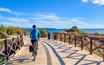 Fietser in blauwe jas op geplaveid kustpad met houten hekken, Middellandse Zee en blauwe lucht zichtbaar op achtergrond.
