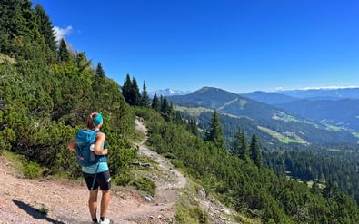 Wandelaar met blauwe rugzak op Hochkönig pad met uitzicht op groene beboste dalen en verre Alpentoppen onder blauwe hemel.