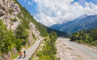 Twee fietsers op geasfalteerd fietspad langs rivierdal met kalksteenrotsen, bos en Alpengebergte onder bewolkte hemel.