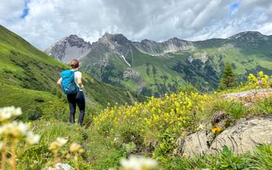 Vrouwelijke wandelaar met blauwe rugzak op groene alpenweide kijkend naar bergpanorama van Steeg naar Landeck met wilde bloemen op voorgrond.