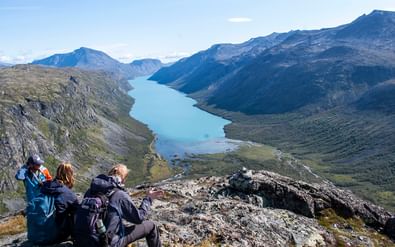 Drie wandelaars met rugzakken zitten op rotsachtige top en kijken uit over turquoise meer omringd door bergen in Jotunheimen, Noorwegen.