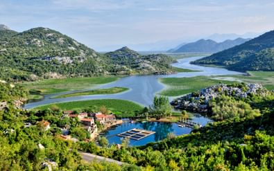 Luchtfoto van het Skutarimeer in Montenegro met kronkelende waterwegen, groene heuvels en een klein dorp met houten steigers aan de oever.