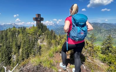 Vrouwelijke wandelaar met blauwe rugzak op bergkam, kijkend naar het grote Jakobskreuz kruis op Buchensteinwand met Alpen op de achtergrond.