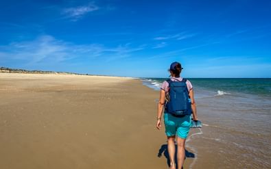 Vrouwelijke wandelaar met blauwe rugzak loopt blootsvoets langs zandstrand met turquoise oceaangolven onder blauwe lucht op Portugese Camino.