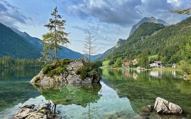 Hintersee meer met kristalhelder water dat bergen en wolken weerkaatst. Rotseiland met bomen in het midden, Alpengebouwen aan de oever.