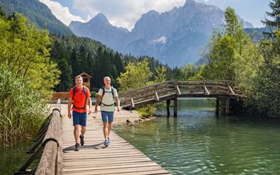 Twee wandelaars met rugzakken lopen over houten loopbrug boven turquoise Jasna meer in Slovenië, met Julische Alpen op achtergrond.
