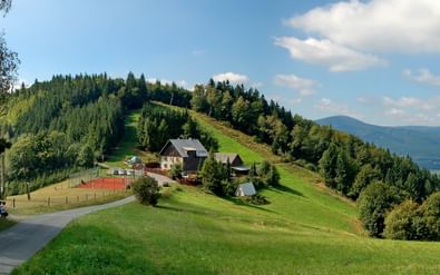 Panoramisch uitzicht op groene glooiende heuvels in Beskydy, Tsjechië met verspreide huizen, een kronkelende weg en beboste hellingen onder een blauwe lucht.