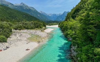 Turquoise Soča-rivier stroomt door Julische Alpen-vallei met witte kiezelstranden, weelderige groene bossen en besneeuwde bergen op achtergrond.