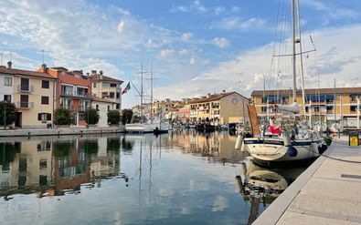 Rustig havenkanaal in Grado met afgemeerde zeilboten, kleurrijke gebouwen langs de waterkant en reflecties in het kalme water onder een blauwe lucht.