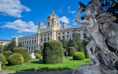 Kunsthistorisches Museum in Wenen met sierlijke koepel en gevel. Verzorgde tuinen met gesnoeide hagen en klassiek standbeeld op voorgrond.
