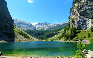 Helder groen bergmeer omringd door steile rotskliffen en bossen in Slovenië's Triglav-regio, met bergtoppen op de achtergrond.