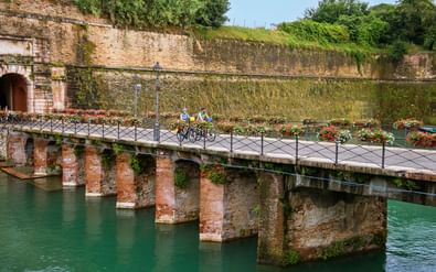 Twee fietsers steken een met bloemen versierde stenen brug over turquoise water over, met oude vestingmuren en groene vegetatie op de achtergrond.