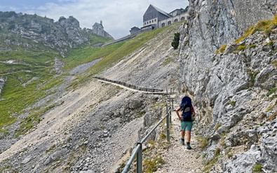 Wandelaarster met rugzak beklimt rotsachtig bergpad op Wendelstein met metalen leuningen. Bergstation zichtbaar op de top.