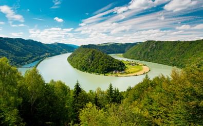 Luchtfoto van de Donau die een hoefijzerbocht vormt rond een beboste heuvel in Oostenrijk, met groene bossen en blauwe lucht.