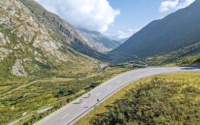 Twee fietsers op kronkelende bergweg bij Lukmanierpass met dramatische Alpenvallei en rotsachtige toppen onder bewolkte hemel.